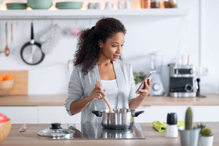 Shot of confident young african american woman cooking healthy food while using her mobile phone in the kitchen at home.の写真素材