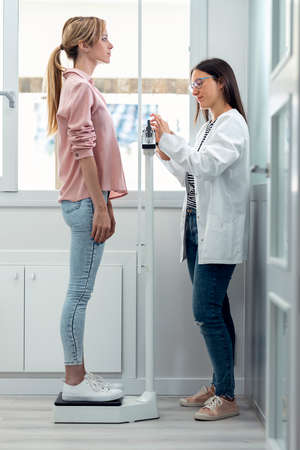 Shot of beautiful young nutritionist woman weighing and measuring his patient on the scale in consultation.の写真素材