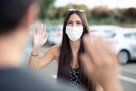 Shot of young beautiful woman student wearing surgical mask and waving hand for greeting to her friend in university campus at Covid19 outbreak. Social distancing and new normal concept.の写真素材
