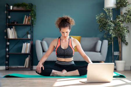 Shot of confident sporty young woman doing hypopressive exercises following online gym classes via laptop on floor in her living room at home.の写真素材