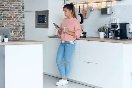 Shot of smiling young woman using her mobile phone while drinking a cup of coffee in the kitchen at home.の写真素材