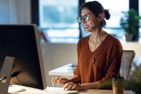 Shot of beautiful young business woman working with computer while talking with earphone sitting in...