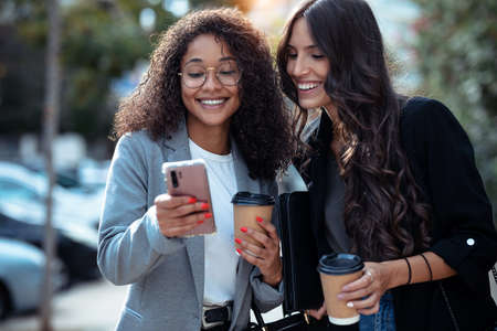 Shot of two beautiful businesswomen using smart phone while drinking coffee walking around the city.の写真素材