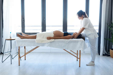 Shot of beautiful young physiotherapist doing osteopathic or chiropractic treatment in pregnant woman's neck on a stretcher at home.の写真素材