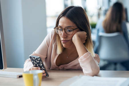Shot of boring young business woman sending messages with mobile phone while sitting in modern...