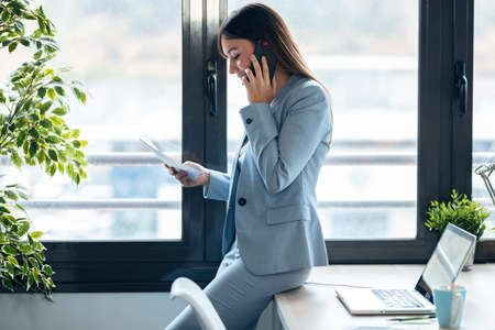 Shot of smart young businesswoman talking with mobile phone while cheeking some documents in modern workspace.の写真素材