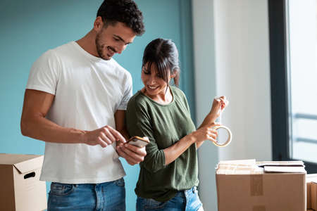 Shot of attractive young couple packing boxes to move to new flat while looking mobile phone in...