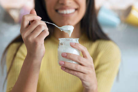 Portrait of pretty young woman looking at camera while eating yogurt sitting on the kitchen at home.の写真素材