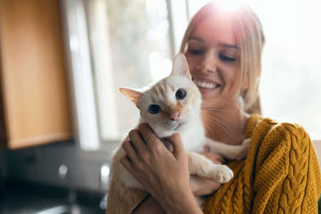 Shot of attractive smiling woman embracing her lovely cat while playing with it in the kitchen at...