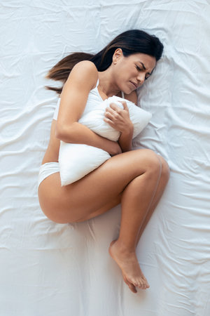 Shot of unhealthy beautiful young woman with stomachache while hugging the pillow lying in the bedroom at home.の写真素材