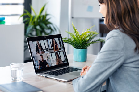 Back view of beautiful young business woman making video call with computer while talking with earphone sitting in modern startup office.の写真素材