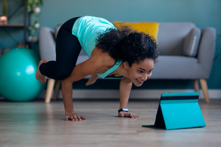 Shot of afro sporty young woman doing hypopressive exercises following online gym classes via...