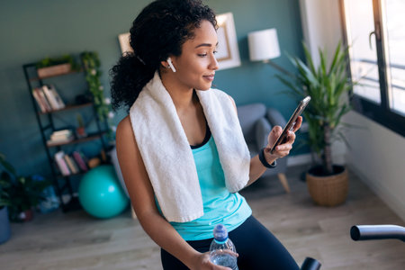 Shot of afro young fitness girl using mobile phone while training on exercise bike at home.