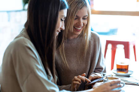 Shot of two beautiful women friends drinking while looking smart phone in the restaurant.の写真素材