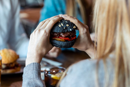 Close up of blonde young woman eating delicious burger while having fun with her friends in the restaurant.の写真素材