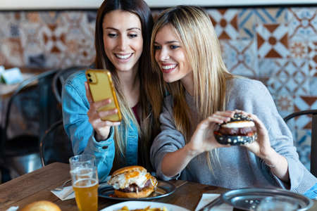 Shot of two beautiful young women friends eating burgers while looking smart phone in the restaurant.の写真素材
