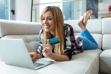 Shot of smiling young woman paying something online with her credit card in laptop while lying on couch at home.の写真素材