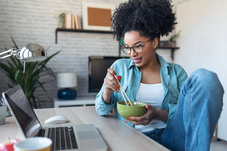 Shot of beautiful young afro business woman making video call with laptop while eating noodles with chopsticks sitting in the office at home.の写真素材