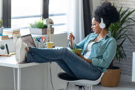 Shot of beautiful young afro business woman making video call with laptop while eating noodles with...