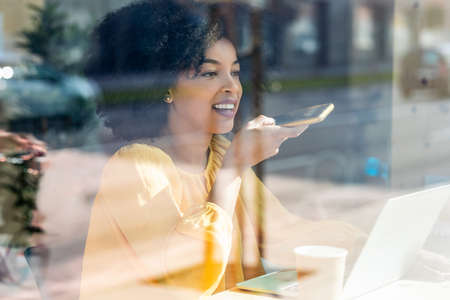 Shot of beautiful afro business woman sending audio voice message with her smartphone while working with laptop in coworking place.の写真素材