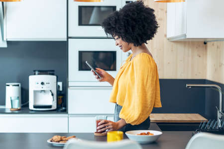Shot of beautiful afro woman sending messages with her smartphone while drinking tea in the kitchen at coworking place.の写真素材