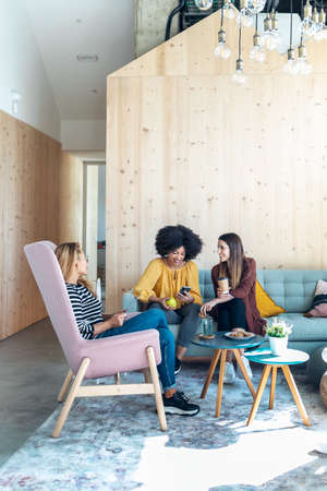 Shot of smart entrepreneur multiethnic women talking while taking a break and showing photos with smart phone sitting on couch at coworking place.の写真素材