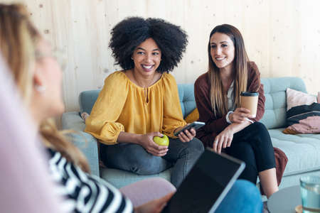 Shot of smart entrepreneur multiethnic women talking while taking a break and showing photos with smart phone sitting on couch at coworking place.の写真素材