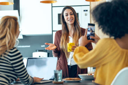 Shot of three smart entrepreneur women talking while taking a break and having a breakfast while taking a photo in the kitchen at coworking place.の写真素材