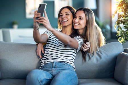 Shot of two happy beautiful women doing a video call with laptop while hugging each other sitting on the couch at home.の写真素材