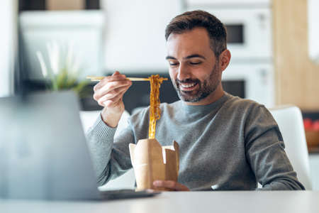 Shot of handsome mature business man eating take away noodles while working with laptop at home.の写真素材