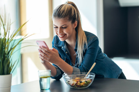 Shot of beautiful young woman eating a bowl of salad while using her mobile phone in the kitchen at home.の写真素材
