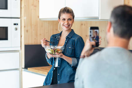 Shot of funny cute couple eating and taking photos with smartphone in the kitchen at home.の写真素材