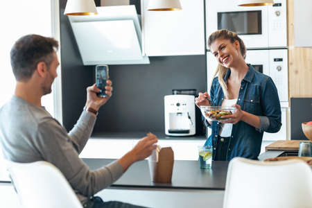 Shot of funny cute couple eating and taking photos with smartphone in the kitchen at home.の写真素材
