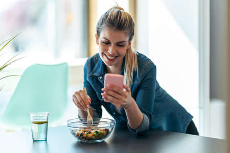 Shot of beautiful young woman eating a bowl of salad while using her mobile phone in the kitchen at home.の写真素材