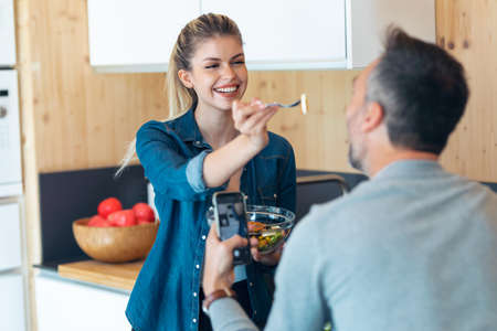 Shot of funny cute couple eating and taking photos with smartphone in the kitchen at home.の写真素材