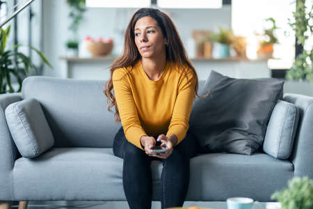 Shot of worried exhausted woman looking her smartphone while waiting for news sitting on couch at...