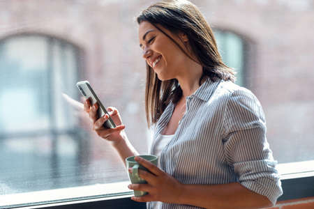 Shot of beautiful young woman using her mobile phone while standing next to the window at home.