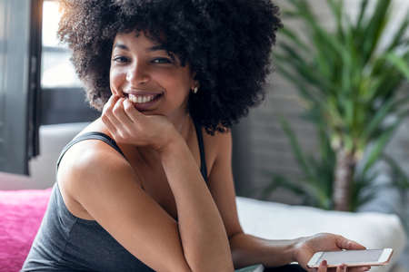 Portrait of beautiful afro young woman using mobile phone while smiling looking at camera at home.