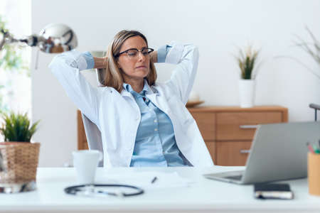 Shot of beautiful mature female doctor stretching body for relaxing while working with laptop in the...