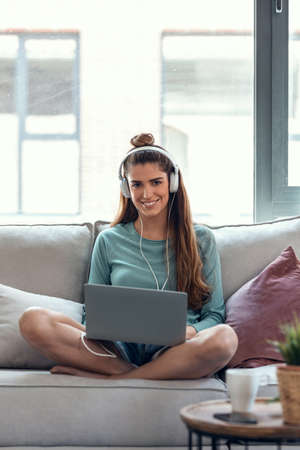 Shot of confident young woman using her laptop while listening to music with headphones sitting on a couch at home.の写真素材