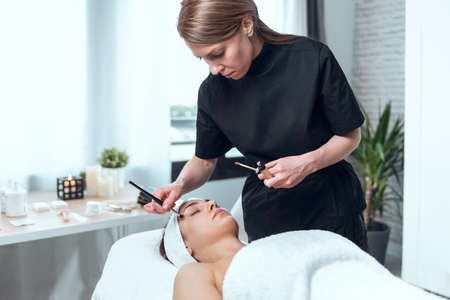 Shot of cosmetologist applying make-up to beautiful woman lying on a stretcher in the spa center.