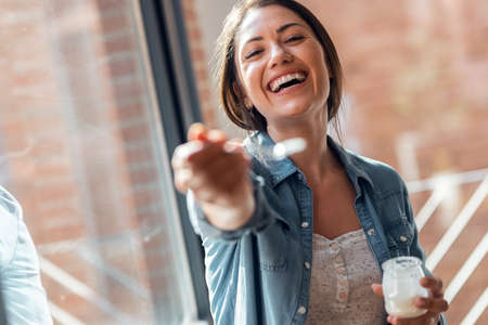 Portrait of beautiful smiling woman having fun while feeding yogurt to camera at home.の写真素材