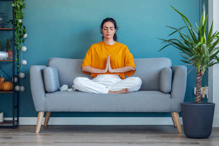 Shot of pretty young woman in lotus position sitting on the sofa at living room at home.の写真素材
