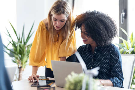 Shot of business women working together with laptop an a digital tablet while talking in modern workspace.の写真素材