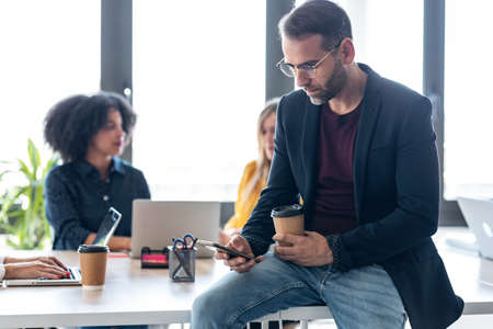 Shot of handosme mature man using her mobile phone while drinking coffee. In the background, her colleagues working in the office.の写真素材