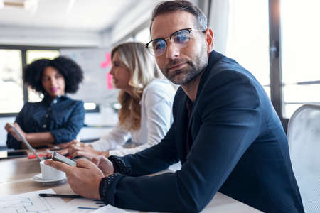 Shot of handosme mature man using her mobile phone in a meeting while looking at camera. In the background, her colleagues working in modern startup.の写真素材