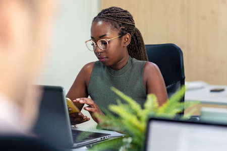 Shot of afro business woman sending messages with mobile phone while working with laptop in a...