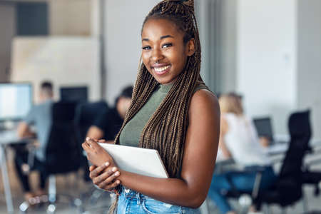 Portrait of smiling young entrepreneur woman using her digital tablet while standing and looking at camera in a coworking place.の写真素材