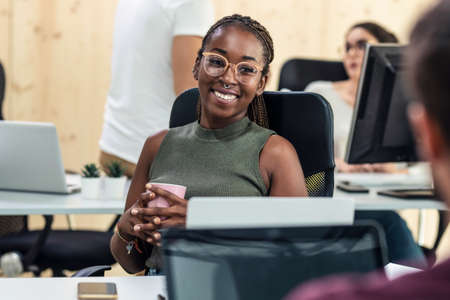 Shot of afro smiling business woman talking with her colleague while working with laptop in a coworking place.の写真素材