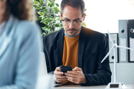 Shot of handsome mature entrepreneur sending messages with smart phone while working with computer in real estate.の写真素材